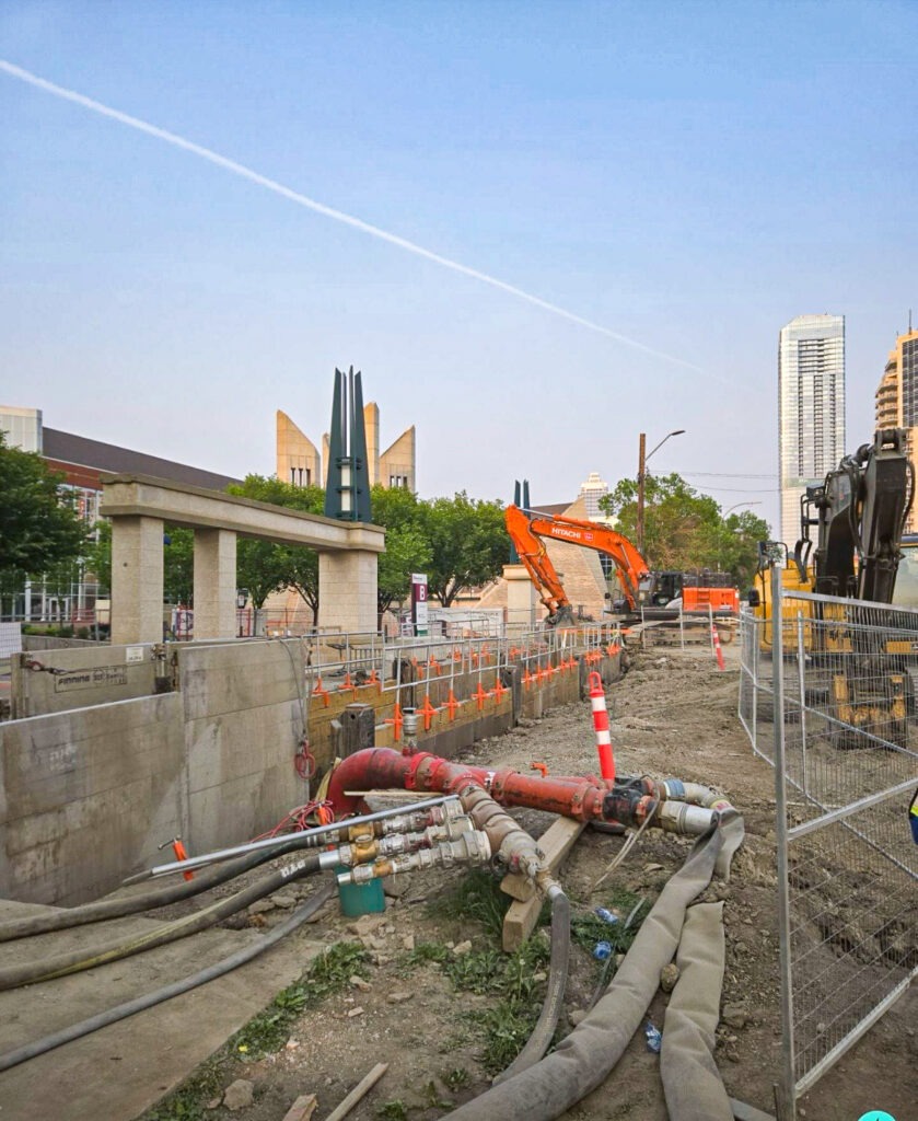 Downtown Edmonton construction site with long shored excavation, heavy equipment, and multiple pipes connected to a large red pipe