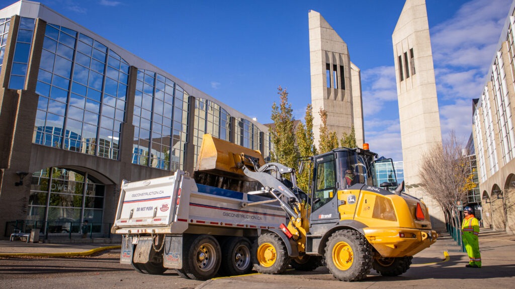 Loader placing soil into a dumper truck while a flagger controls site access